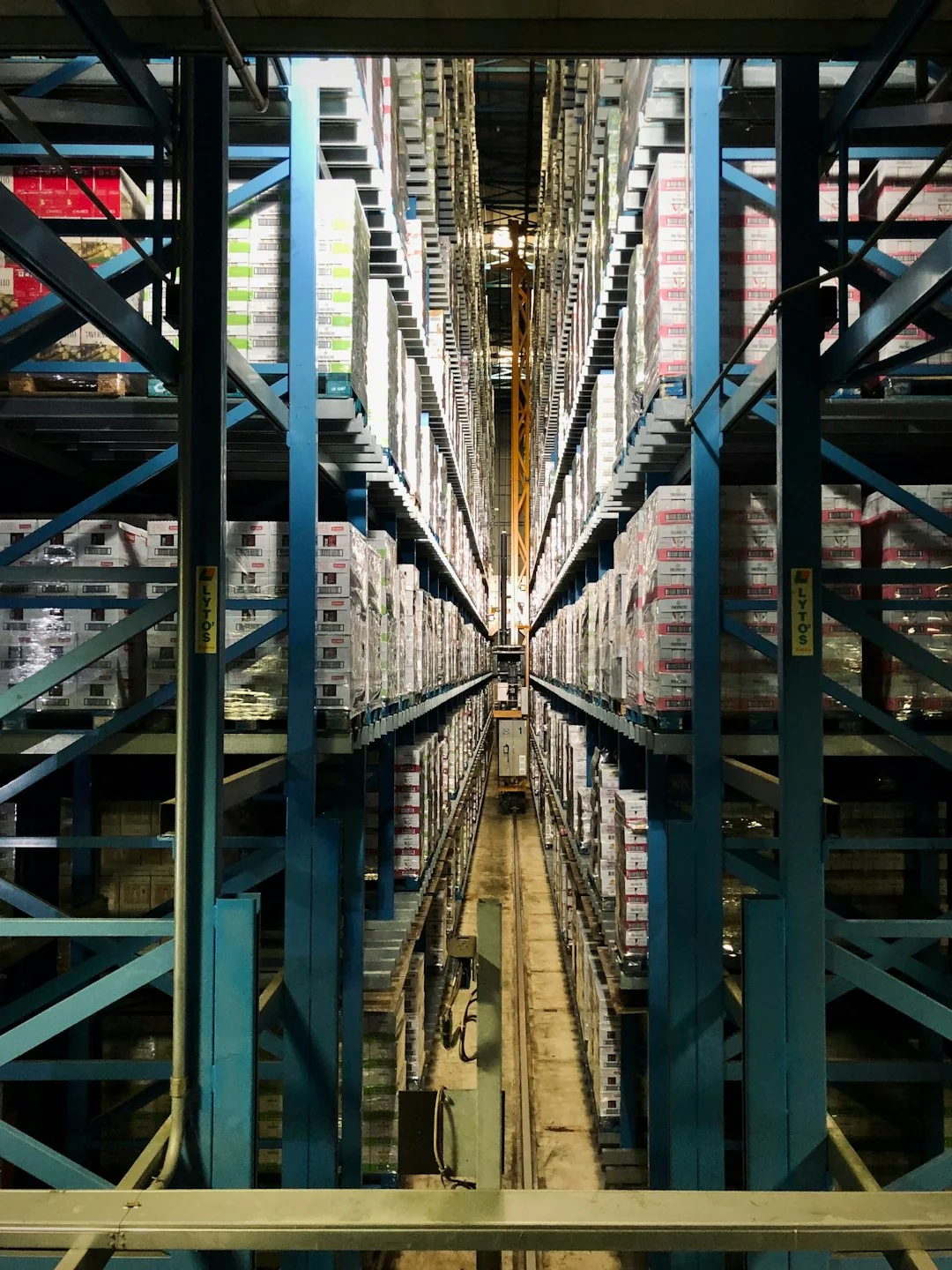 A narrow aisle between blue steel racking stacked to the roof with palletised cartons
